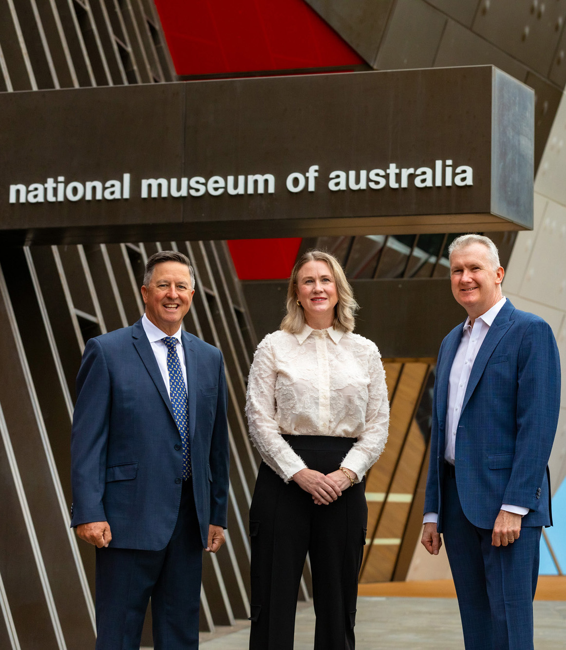 Three people stand outside the front entrance to the National Museum of Australia.