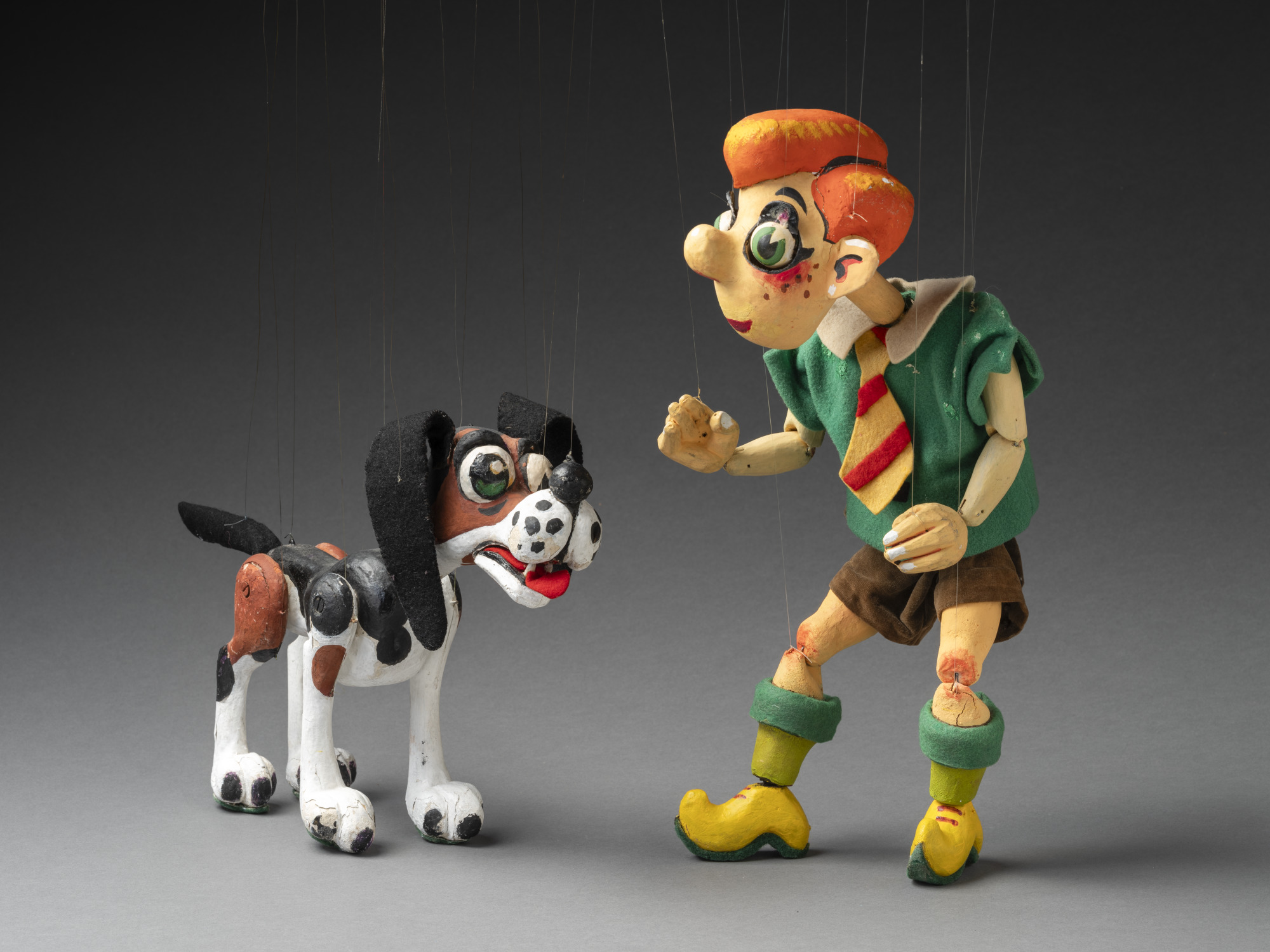 A photo of two string puppets. Right is a puppet of a boy named Nicky, he has bright orange hair and is wearing a green short sleeve shirt, brown shorts and a red and yellow tie. On his legs are green socks and yellow clog-like shoes. Left is a wooden puppet of a dog called Noodle. Noodle has a black, brown and white painted body, felt ears and green felt soles attached to its feet. Noodle has green painted eyes and a red felt tongue.