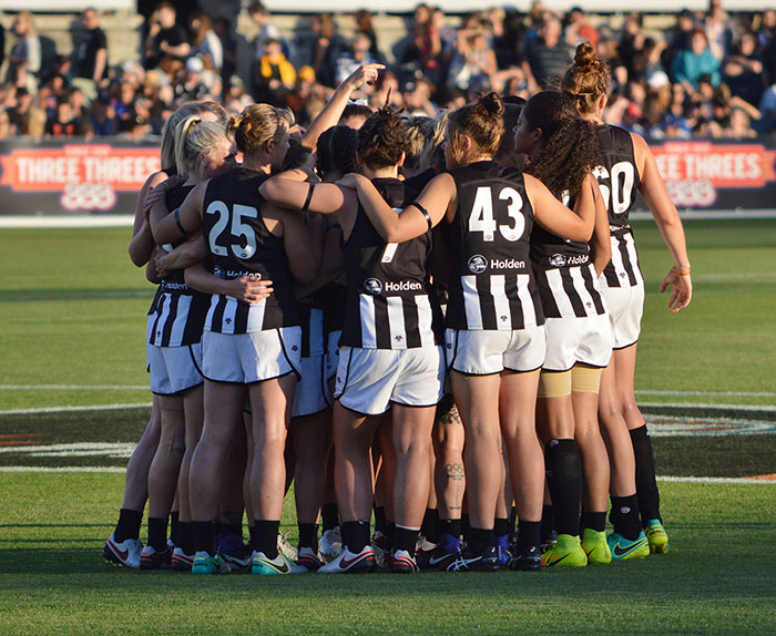 Group of football players wearing black and white stripped Collingwood jerseys and white shorts, forming a circle, with arms around each other, on a field. - click to view larger image