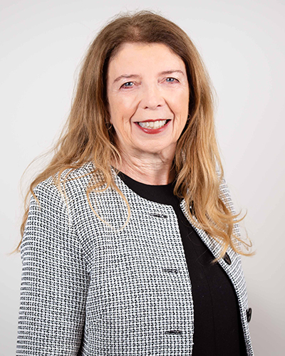 A photograph of Ann McGrath. Ann is wearing a black and white checked blazer and a black shirt. Her hair is strawberry blonde and she is smiling at the camera.