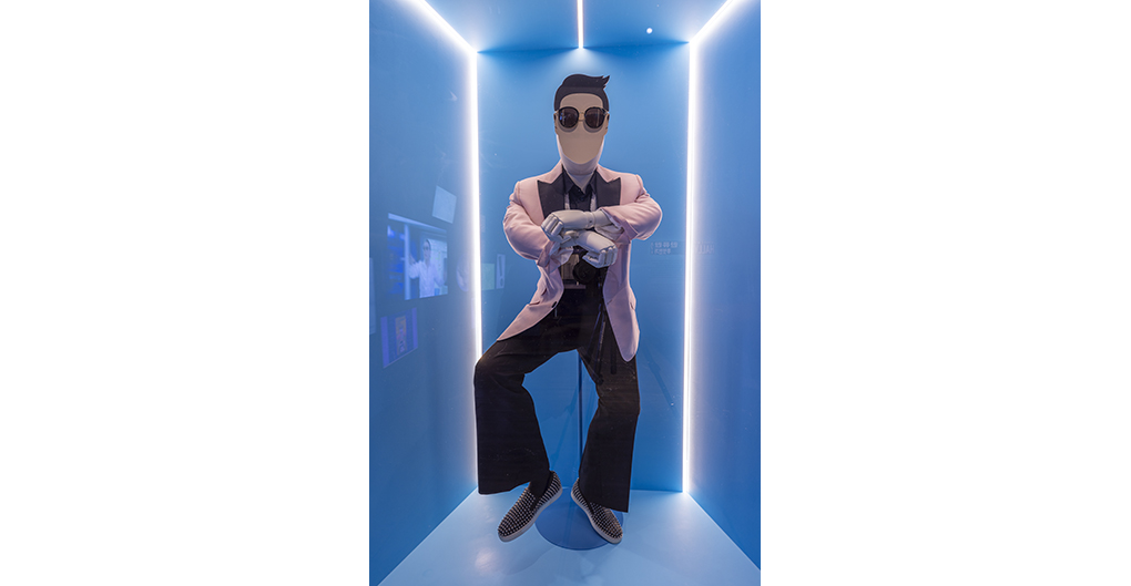 A white mannequin positioned in one of PSY's signature horseriding dance moves. It wears a light pink suit blazer with wide black lapels over a black open collared dress shirt with and around the neck hangs a loose /untied white beaded bowtie. A cardboard mask is attached to the mannequin's face to represent PSY's face. It is featureless save for his slick, black hair style and signature dark sunglasses. THe ensemble is completed with a pair of black trousers and black and white loafers covered with silver studs.