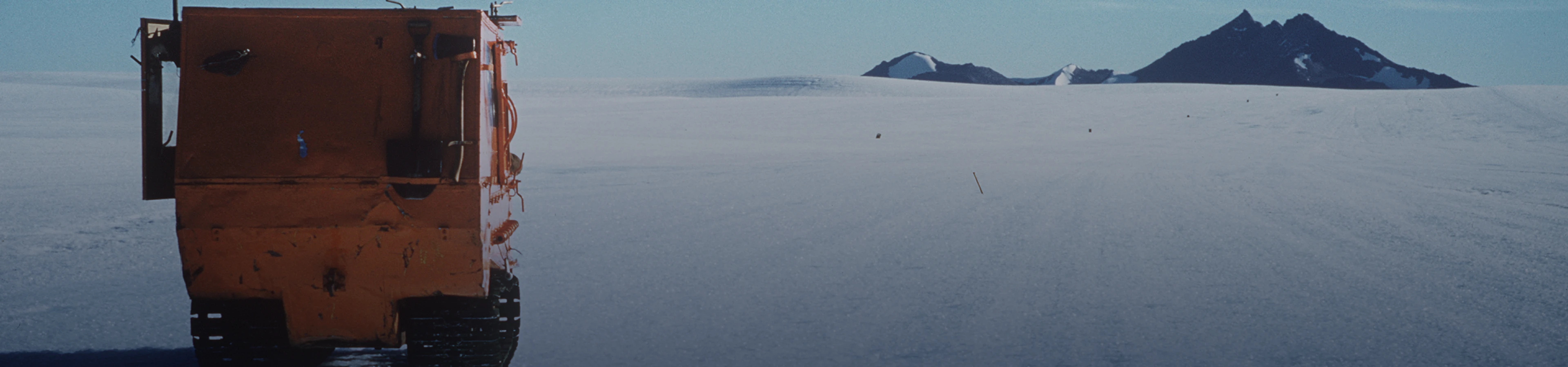 Rear view of a bright orange tracked vehicle in a vast, icy landscape, with rocky mountains on the horizon.