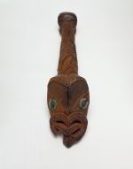 A canoe ornament made of wood and coloured red. The carving features a human face with tongue extended and the larger face with inlaid mother-of -pearl eyes.