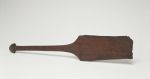 Dance paddle made of wood with an almost rectangular and rounded blade (that is damaged at the lower end) and a semi-circular knob at the end of the handle.