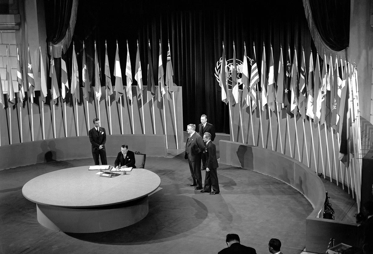 Interior black and white photo of man sitting at large round table signing a document, with four men standing behind him and a series of flags arranged behind them.