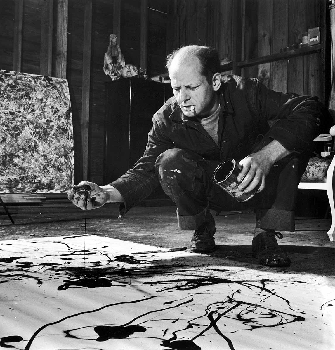 Black and white photo of balding, middle-aged man crouches by canvas spread on the floor. He has a cigarette in his mouth, a paint can in one hand. His other hand seems to be covered in paint. He is frowning intently at the canvas, which has a few splotches of dark paint on it. - click to view larger image