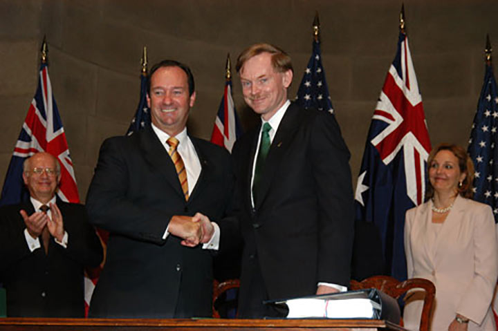 Vaile and Zoellick pose for the cameras shaking hands and smiling. Behind them are a series of Australian and US flags along with a man and a woman smiling and clapping - click to view larger image