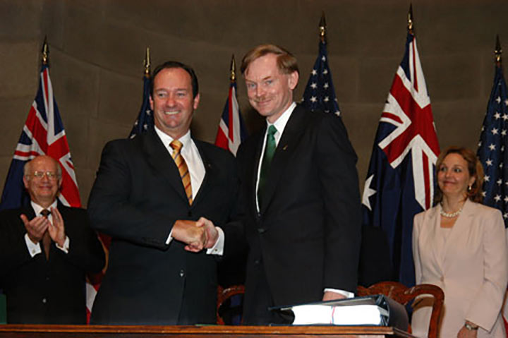 Vaile and Zoellick pose for the cameras shaking hands and smiling. Behind them are a series of Australian and US flags along with a man and a woman smiling and clapping. - click to view larger image