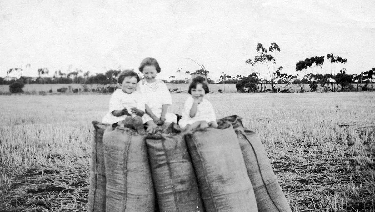 Black and white photograph of three young girls sitting on top of large sacks in paddock.