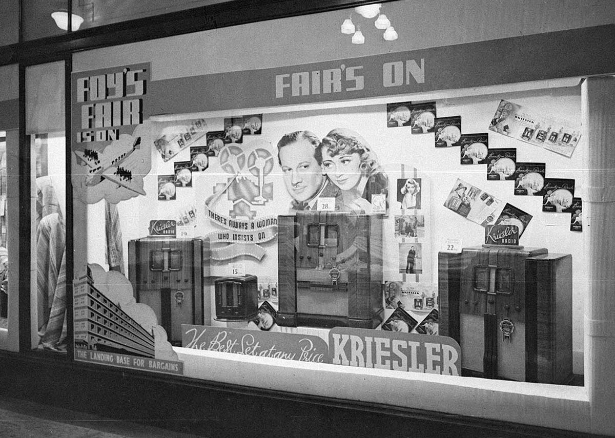 Black and white photograph of a shop front display with radios as the centre peices. They are against a backdrop of various images including a couple, a plane and a building.