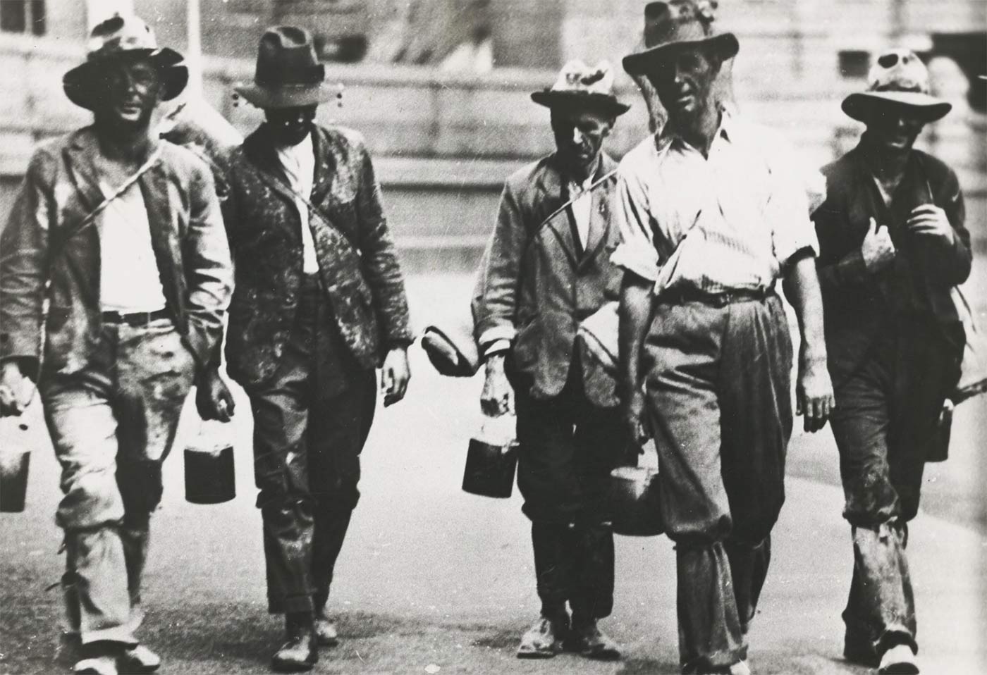 Five men walking towards the camera dressed in shabby clothes. Most are carrying swags and bill cans