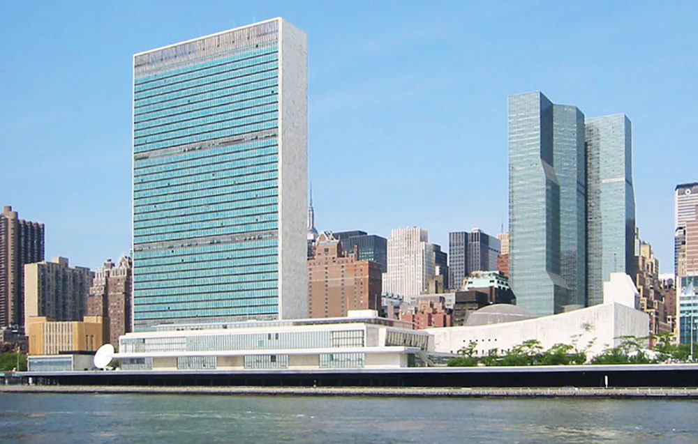 Large glass and concrete building on bank of river, with other buildings on either side.