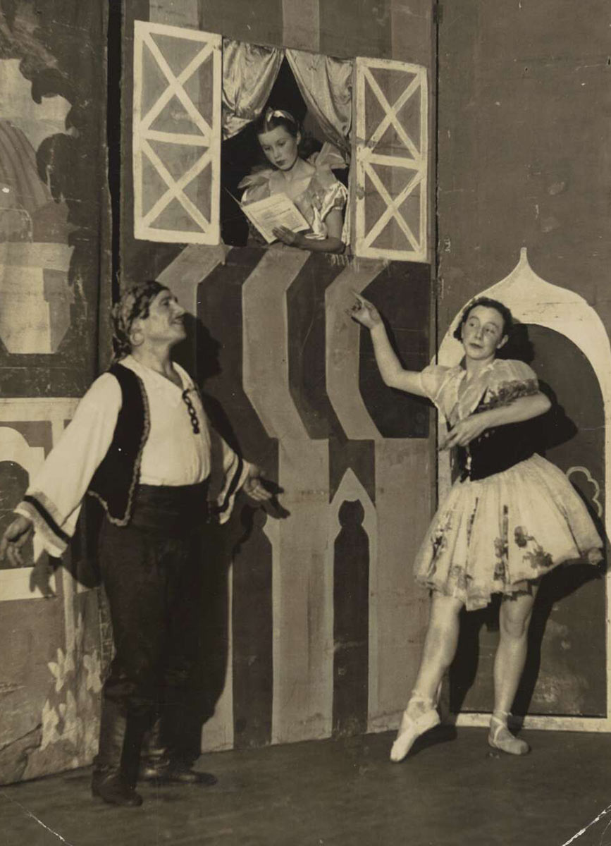 Three performers on stage. Two of them, a man and a woman in traditional folk costume, strike balletic poses while another performer reads a book in a window above their heads - click to view larger image