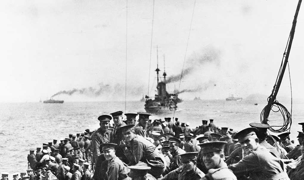 Grainy black and white photo taken on board a boat full of Australian soldiers, with ships in background