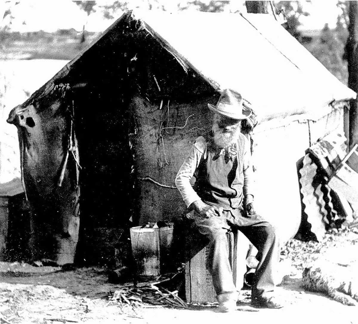 Old man seated on a crate in front of his simple dwelling with a dog at his feet. - click to view larger image