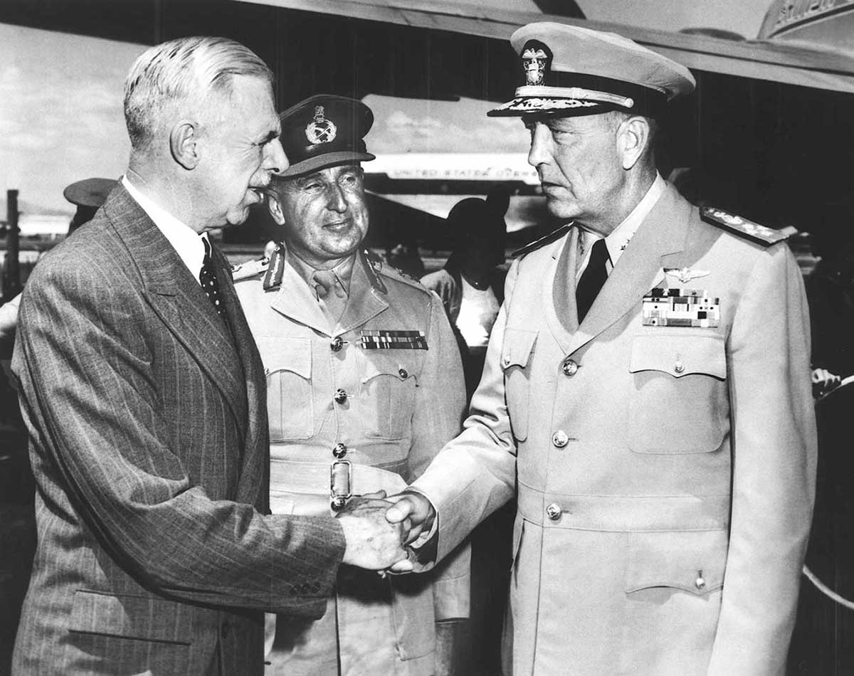 A man in suit shakes hands with US admiral in uniform while New Zealand general looks on.