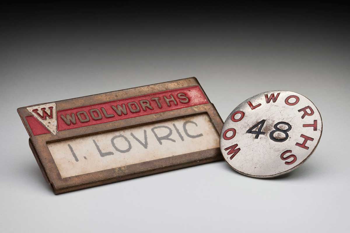 Metal Woolworths name badge printed with ‘I. LOVRIC’, and pin with the number ’48’. - click to view larger image