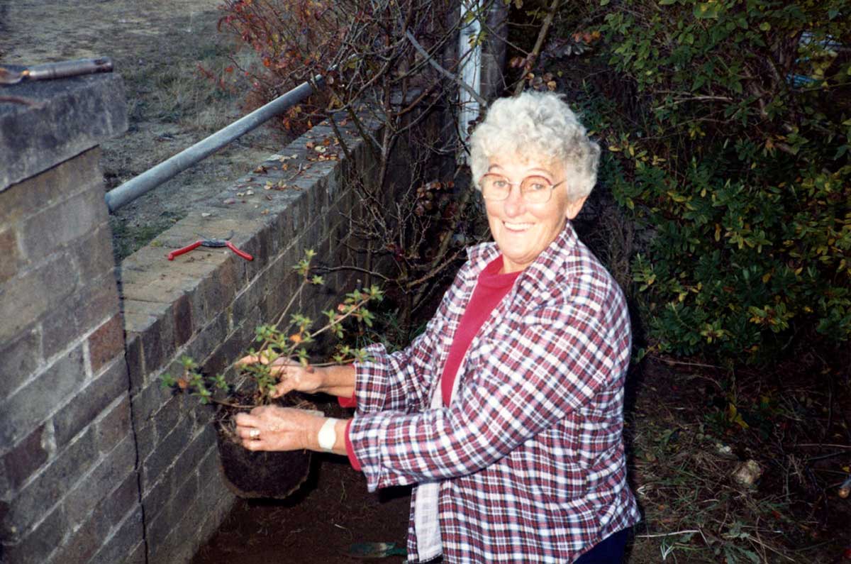 An elderly woman is gardening and smiling towards the camera.