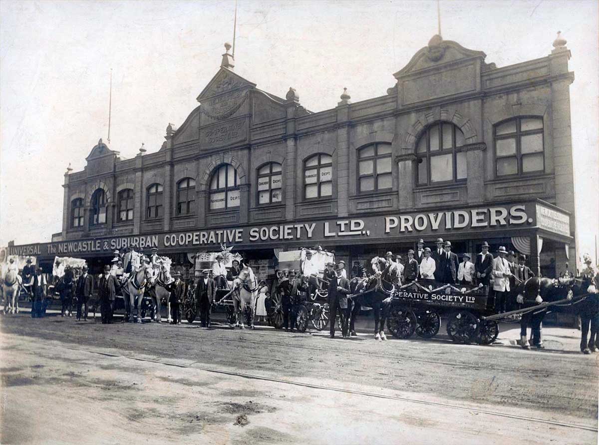 Horse-drawn vehicles lined up at a large shop front.