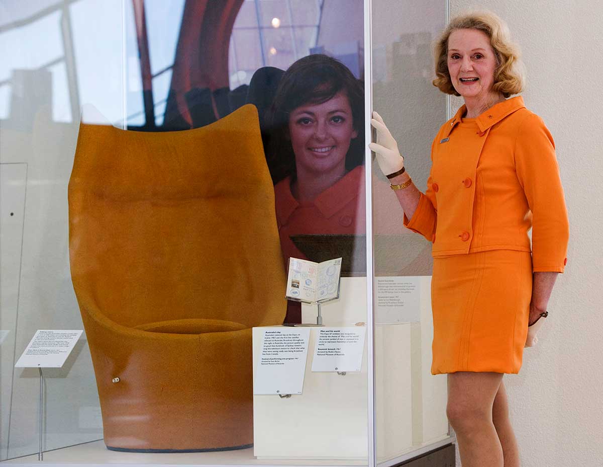 A woman wearing an orange top and skirt stands with her gloved right hand resting on a display case. An orange chair sits in the case, which includes a portrait of another woman at the rear.