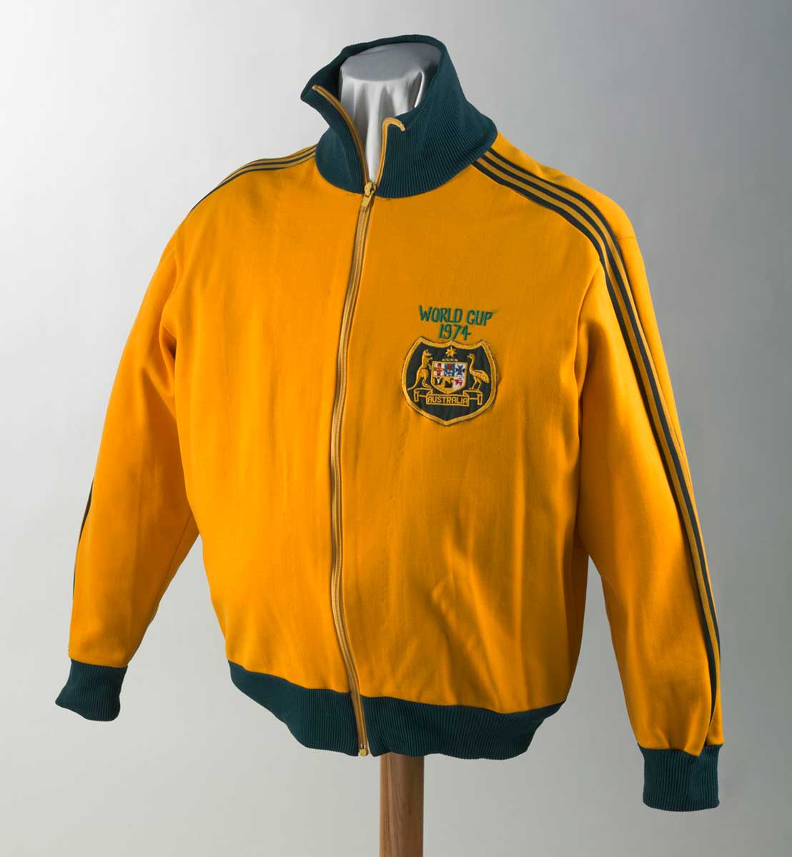 Long-sleeved yellow tracksuit jacket with high elasticated collar, waist and cuffs with zip fastener at front. Australian coat of arms badge sewn front, upper proper left with text above 'WORLD CUP / 1974'. Green stripe detail from shoulder to cuffs along sleeves. - click to view larger image