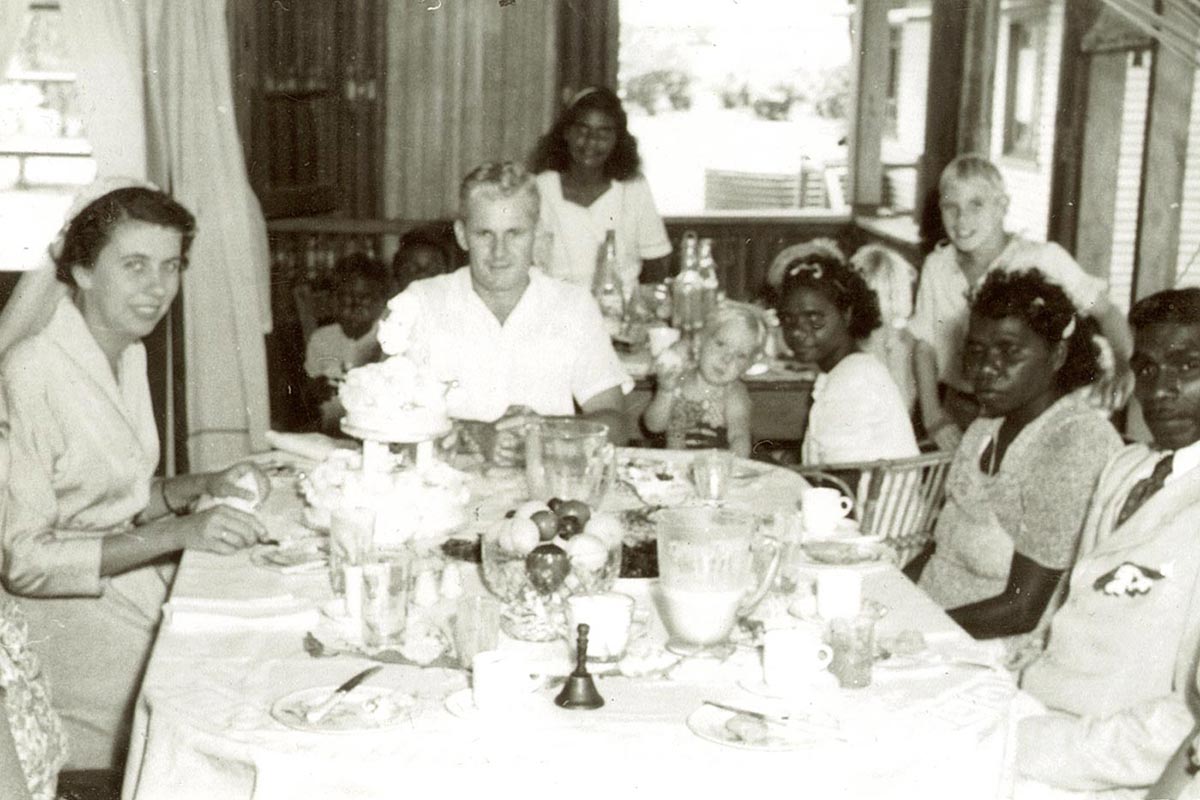 Group of adults sitting at a large tble in the foreground, with children seated at a smaller table behind.