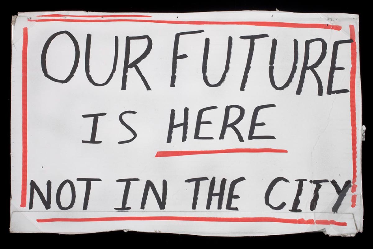 Photograph of a handmade sign on a white piece of card, with a border of red. Black handwritten text reads 'OUR FUTURE IS HERE NOT IN THE CITY'. - click to view larger image