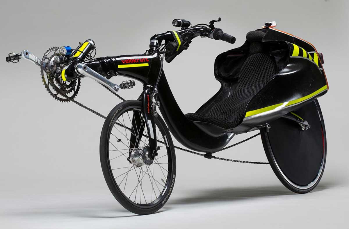A recumbent bicycle with carbon fibre tailbox and two different size wheels. The rear wheel is larger and and has a black wheel cover. The pedals are in front of the front wheel. The steering is above seat. The body of the bicycle is black in colour with some bright yellow stripes. The text ' Velokraft ' is orange in colour and is printed on the frame in front of the bike. The orange colour is also on the top of the tailbox.