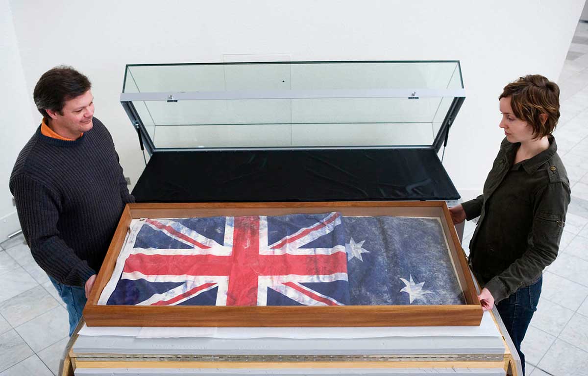 Two Museum staff members moving an Australian flag, retrieved from the ruins of the World Trade Center, into a display case.