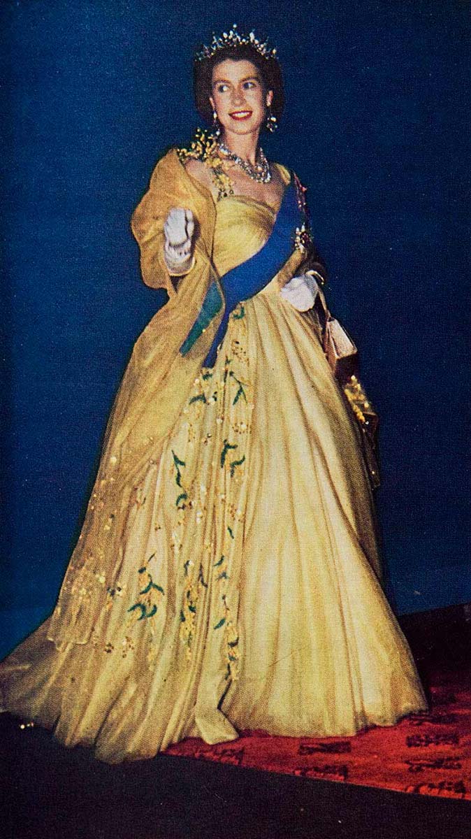 Queen Elizabeth II wearing a floor-length pale-yellow gown, crown, white gloves and blue ceremonal sash. The image shows the queen standing on a red printed carpet, with a deep blue backdrop. - click to view larger image