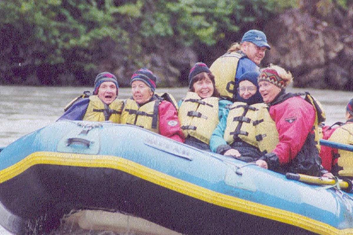 Colour photograph showing six people in an inflatable raft on a river. Five people wearing life jackets sit facing the front of the raft. A man with a ponytail sits higher, towards the rear of the raft. A seventh person is partially visible on the right. - click to view larger image