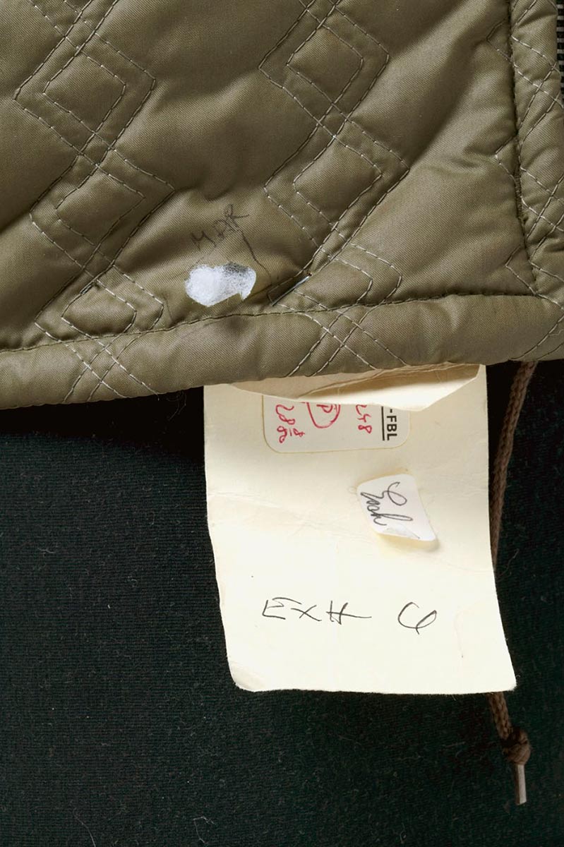 Close up shot of bottom of khaki child's parka, with a paper exhibition tag attached and a section of fabric cut away. - click to view larger image