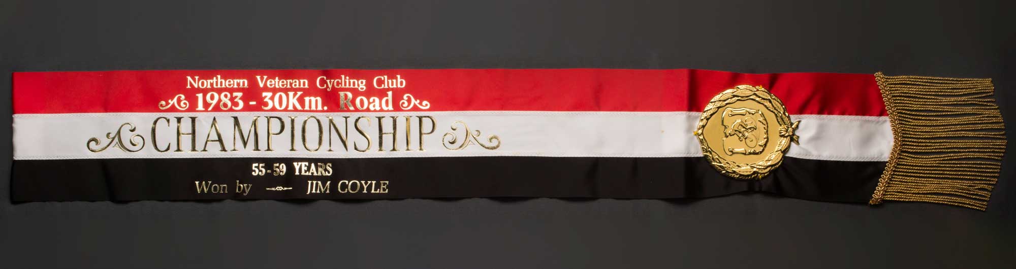 A red, white and black sash made of three ribbons sewn horizontally together. There is gold coloured lettering pressed across the sash. The text reads 'Northern.Veteran.Cycling.Club. / 1983 ? 30Km. Road / CHAMPIONSHIP / 55-59 YEARS / Won by JIM COYLE'. Attached to one side of the sash is a circular shaped medallion with a raised relief of a wreath and a cyclist. The edge of the sash has a gold coloured fringe. - click to view larger image