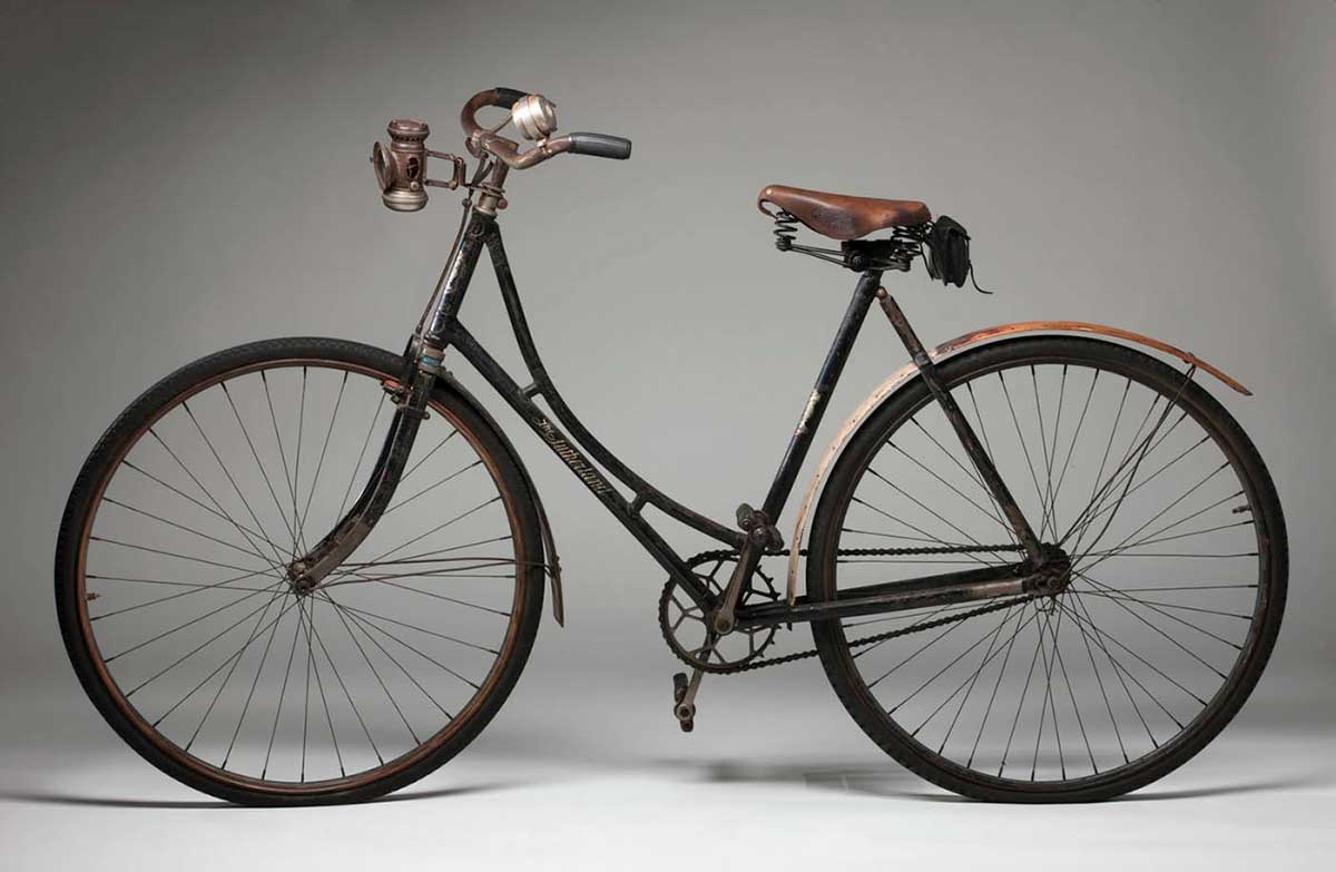 A ladies' bicycle with a black painted metal frame, featuring a painted gold design on the front stem which reads 'The / SUTHERLAND / CYCLE / & MOTOR / WORKS / OPPOSITE / TOORAK STATION / MELBO[UR]NE. It has an oil lamp and a bell attached to the front handle bars and a tool/repair kit attached under the back of the seat. It has wooden mudguards, a steering lock for the front wheel, a front stirrup brake, a rear hand brake and a leather saddle (seat) made by 'BROOKS'. A skip tooth chain wheel and pins hold the bottom bracket cups in position.