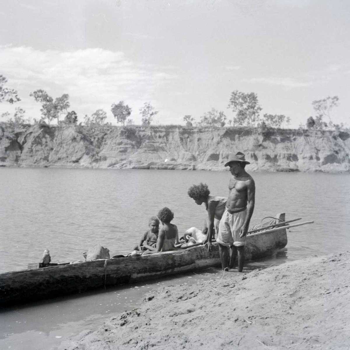 A black and white photographic negative that depicts an Aboriginal man and woman standing on a riverbank next to a canoe with a child and adult sitting in the canoe. - click to view larger image