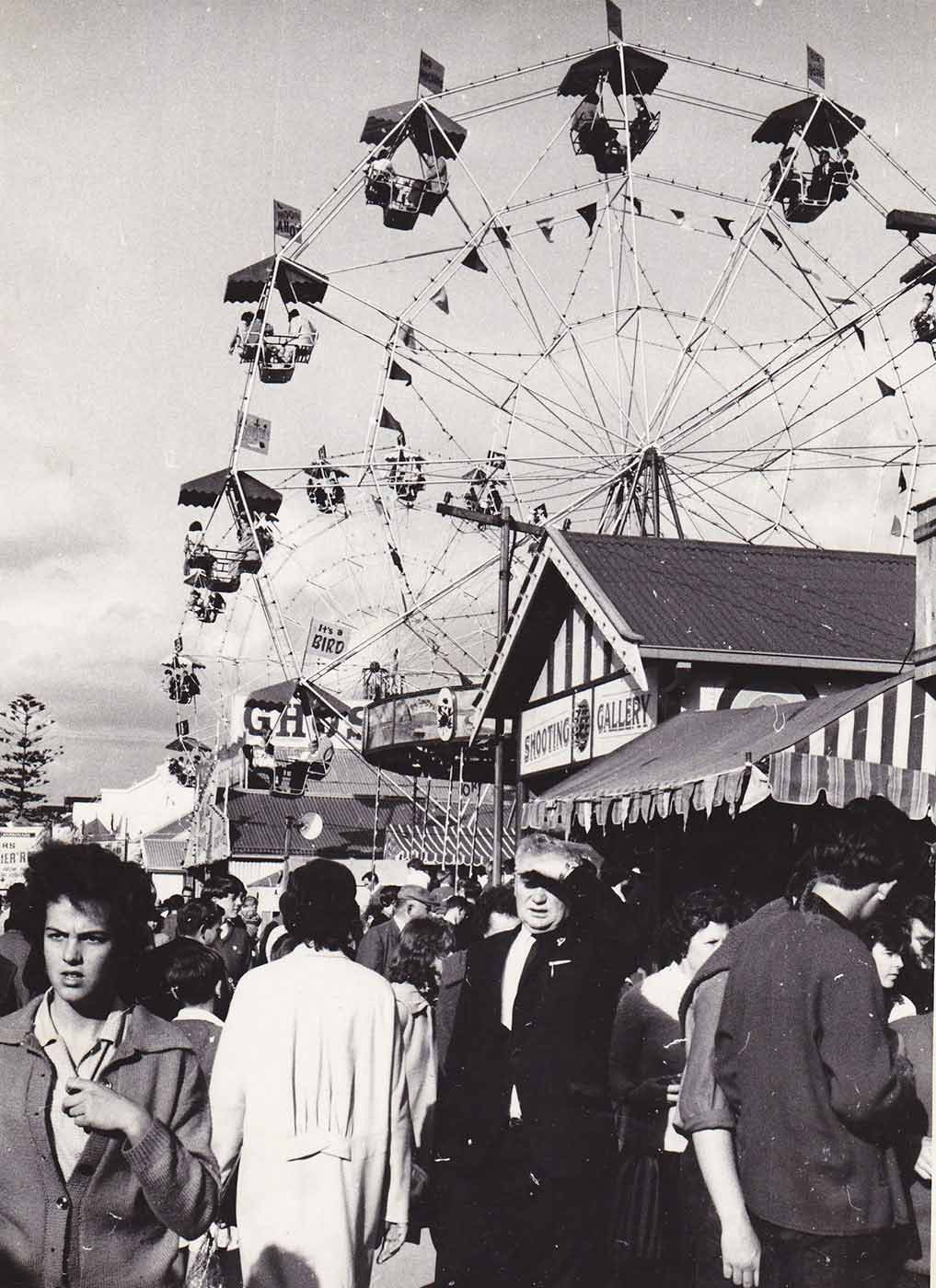 Black and white photo showing a ferris wheel in the distance and a group of people walking down sideshow alley in the foreground. - click to view larger image