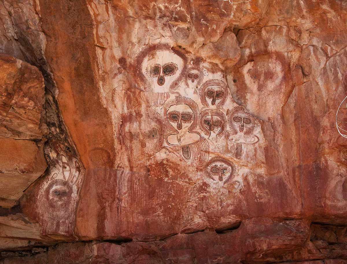 Colour photograph showing Aboriginal art on a rock wall. Numerous faces with oversized eyes and no mouth are depicted on the rock, in shades of yellow, red, black and white. - click to view larger image