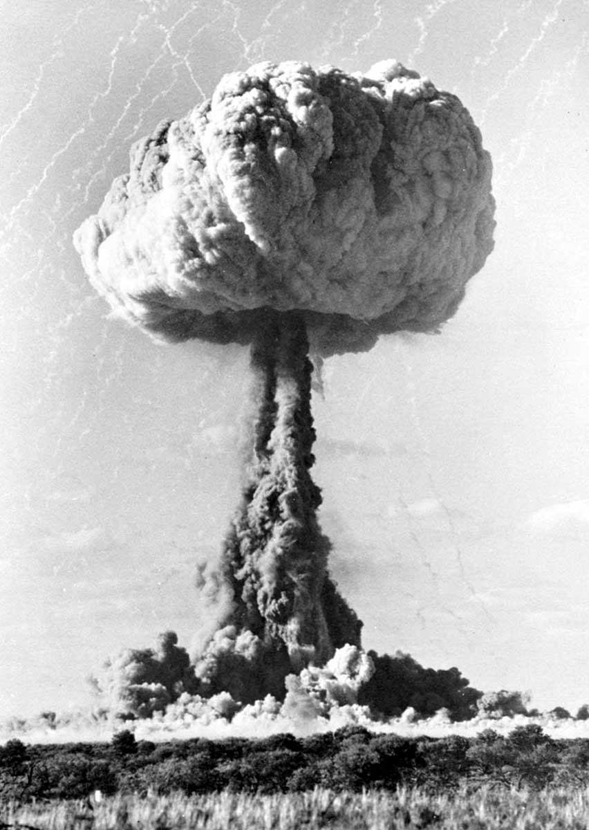 Classic mushroom cloud of a nuclear explosion in the background, with grassy scrub in foreground. - click to view larger image