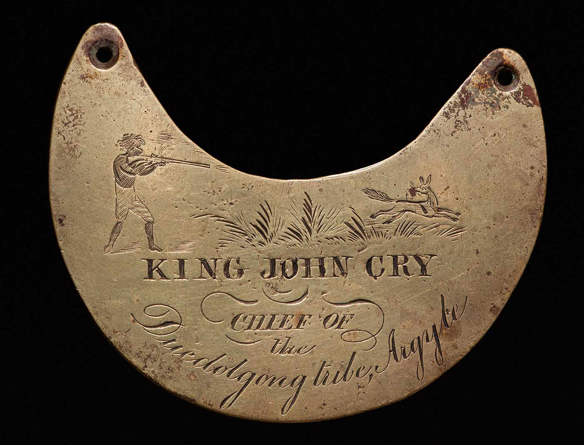 A crescent shaped, brass breastplate with a hole at the edge of each horn to attach a chain. ‘KING JOHN CRY / CHIEF OF / the / Duedolgong Tribe, Argyle’ is engraved on the anterior surface. A man with a rifle is engraved on the left horn, and a fox is engraved on the right horn - click to view larger image