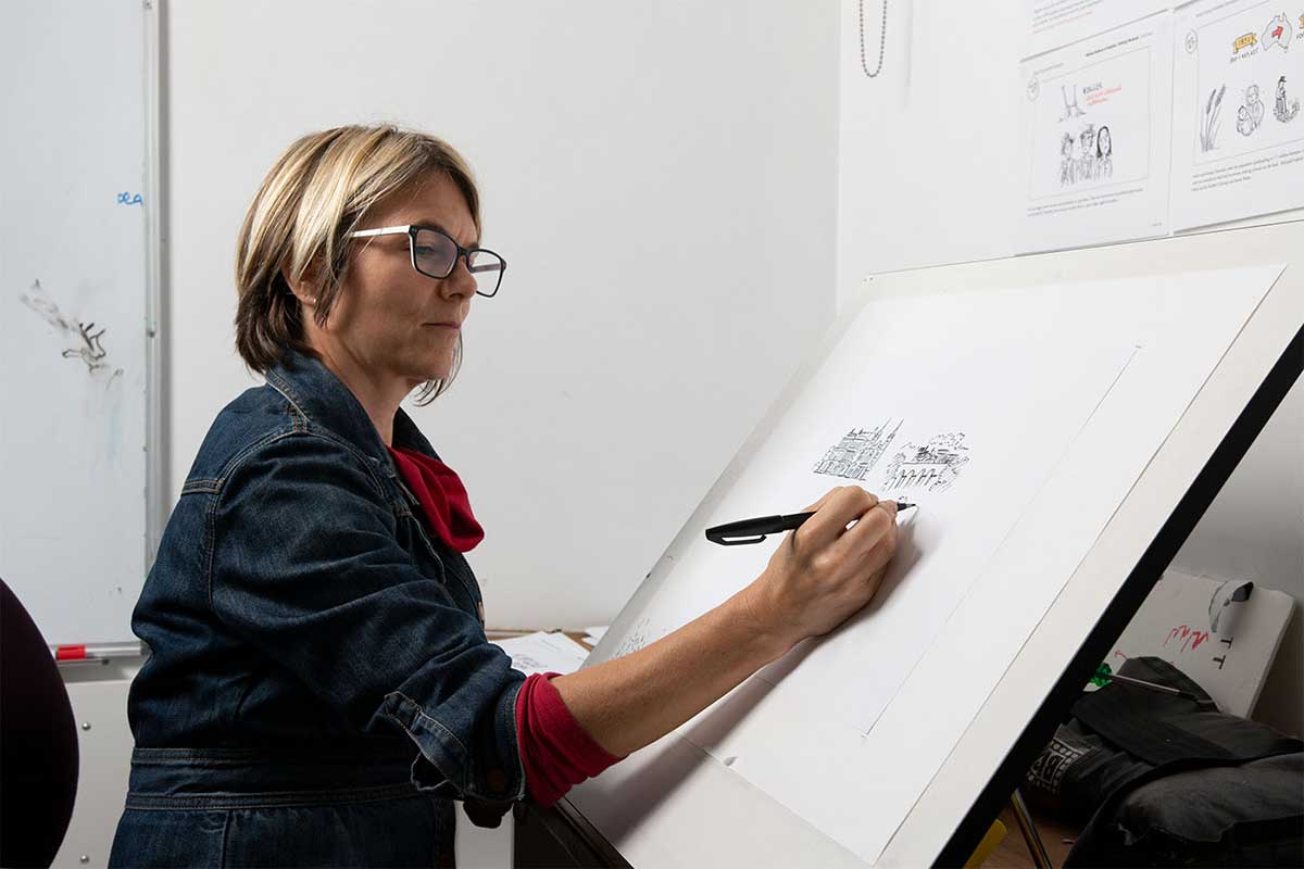 Side view of a woman seated before a large easel, sketching a cityscape on white paper in black pen. - click to view larger image