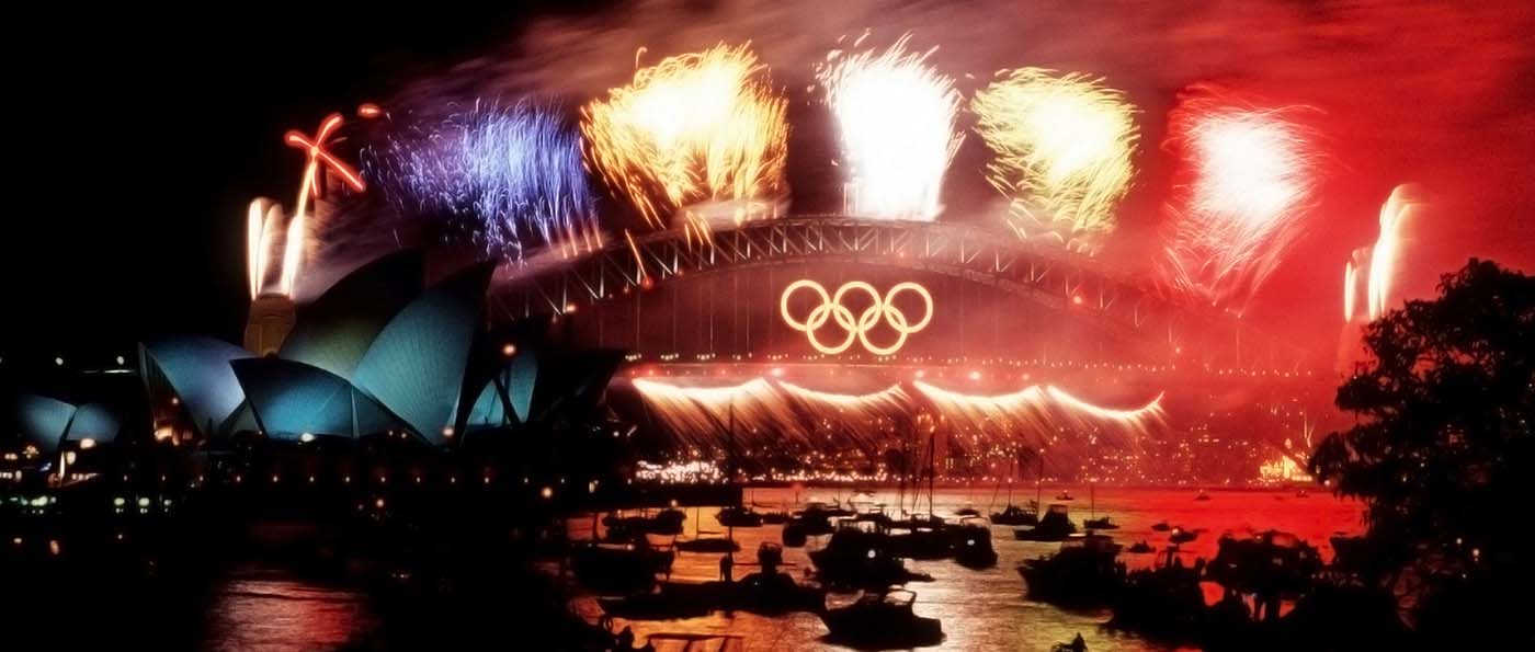 A photo taken at night featuring fireworks on a bridge which has the 5 interlaced Olympic rings centred on the side. The harbour is filled with boats and surrounded by lights in the distance.