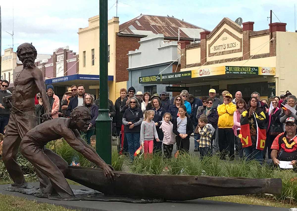 Sculpture of two muscular Aboriginal men with bark canoe. A crowd of people looks on, with shops in the background.