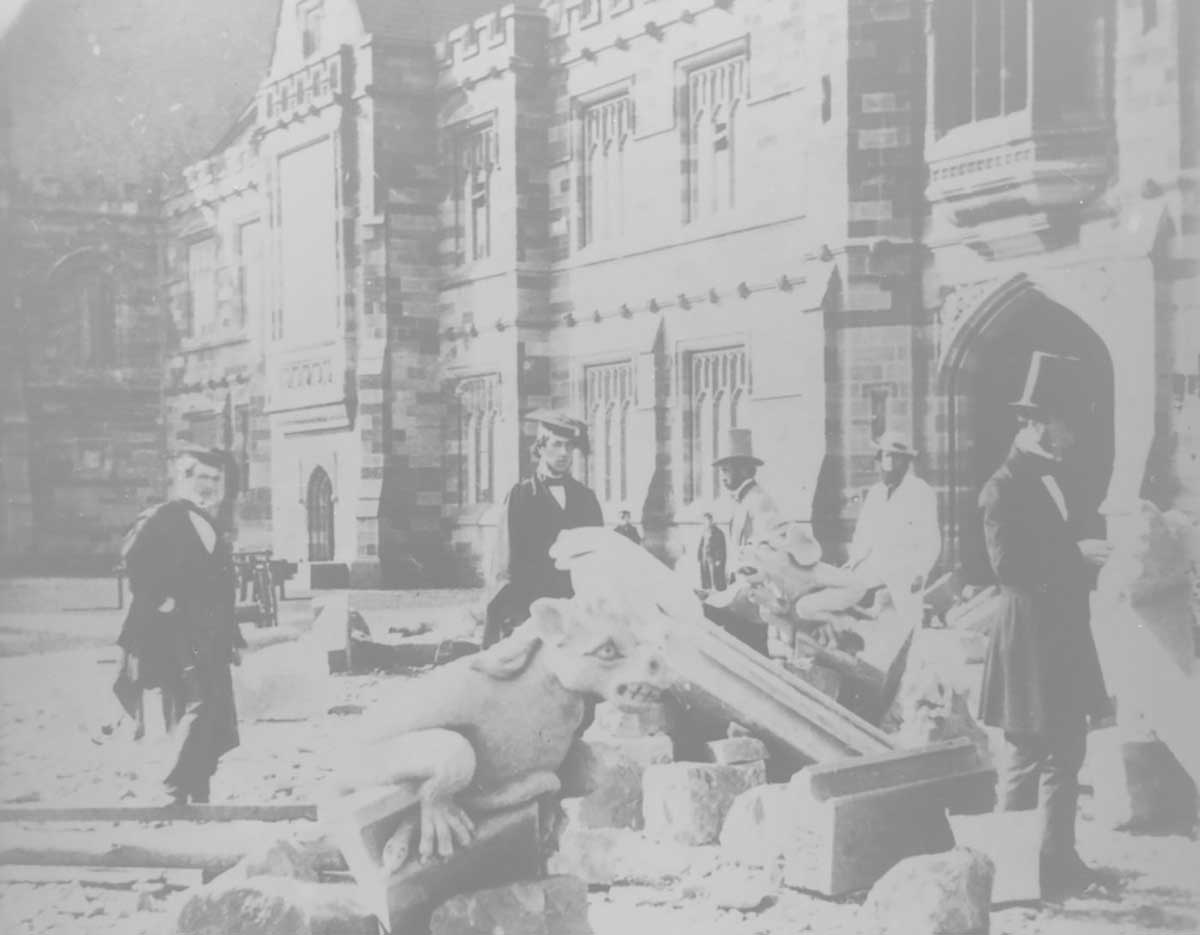 Faded black and white photograph of men amongst rubble, in front of a sandstone building. - click to view larger image