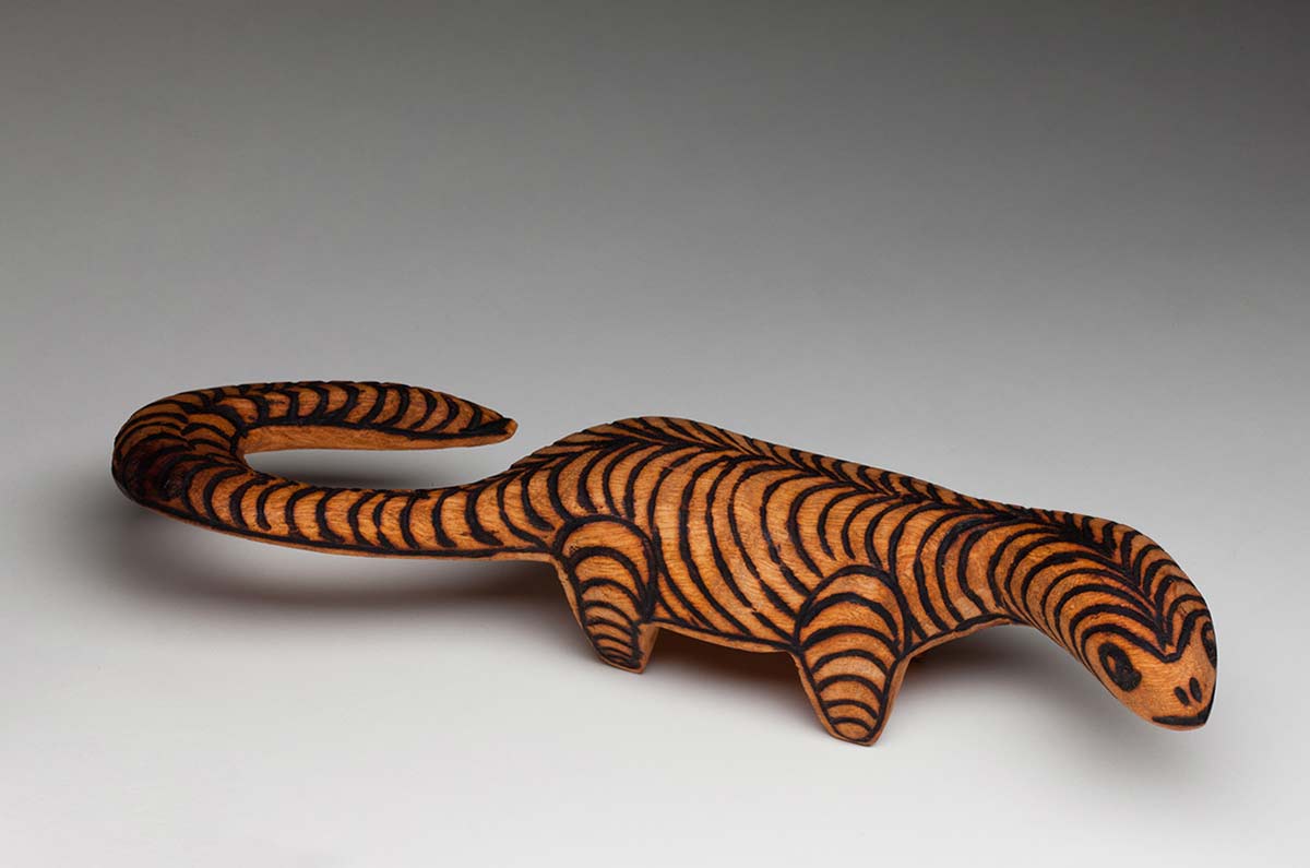 Wooden perente lizard or goanna standing upright on four short legs with its elliptical head turned to the right and a long hooked curled tail. The top exterior is covered with burnt pokerwork designs forming two rows of curving arcs meeting in the centre of the spine while the legs are covered with parallel curved lines. - click to view larger image