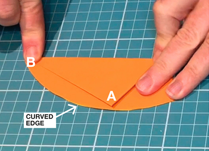 Fingers hold down a piece of orange paper. The letter B in white font is superimposed on the top left corner of the paper and the capital letter A (also in white font) is superimposed on a pointy end in the centre. A few centimetres to the left is a small block of black text in capital letters which reads 'curved edge' with an arrow pointing to the curved edge of the paper.