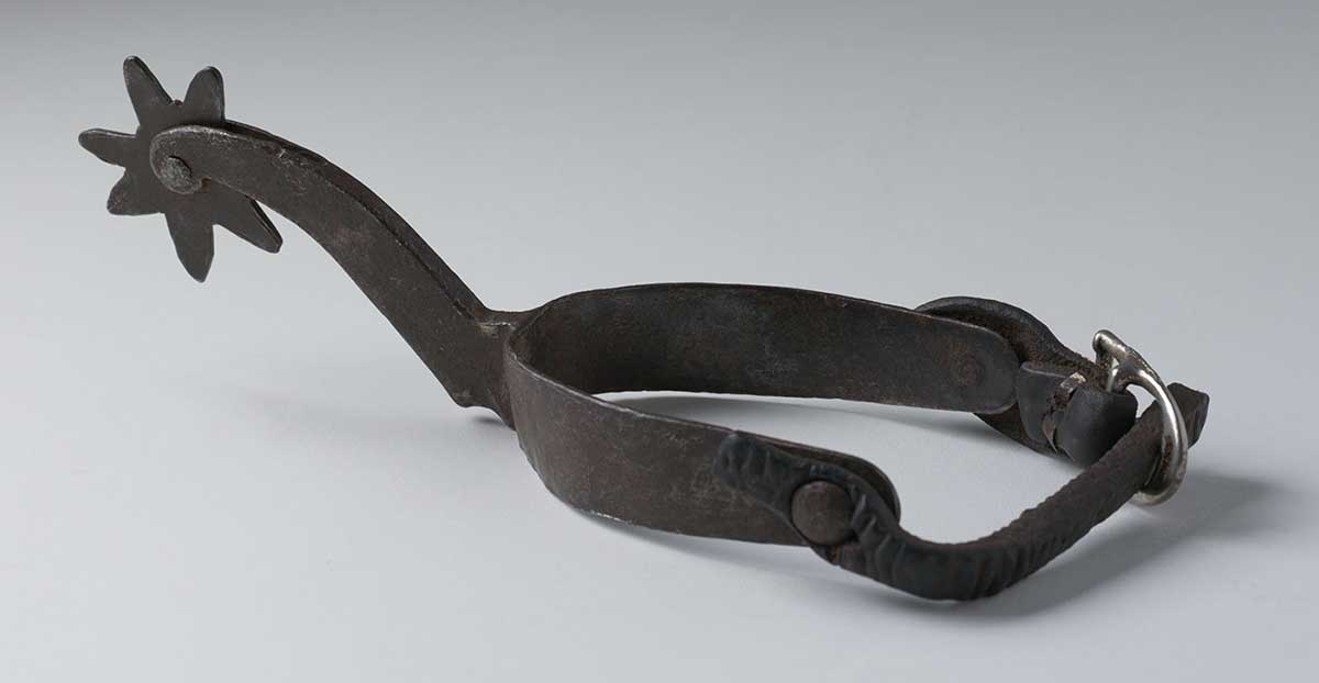 A dark brown metal spur with leather and buckle boot attachment. - click to view larger image