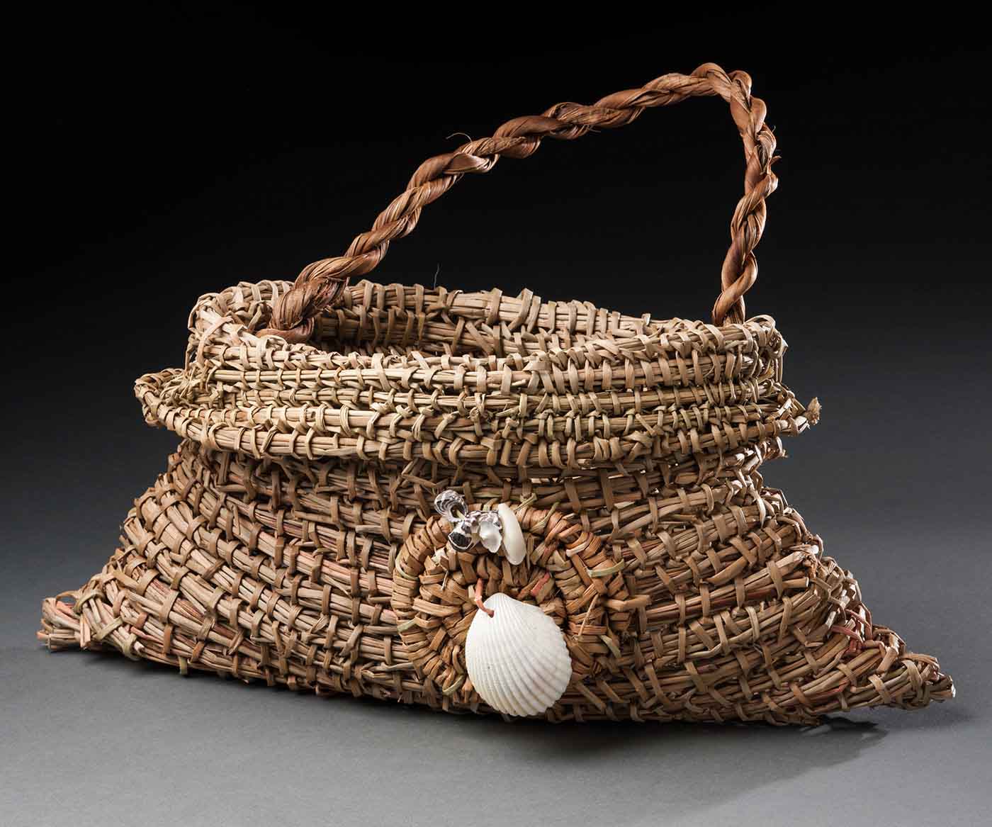A woven bag with a short plaited handle. On the front side of the bag is a decorative coiled circle with five shells attached to it.