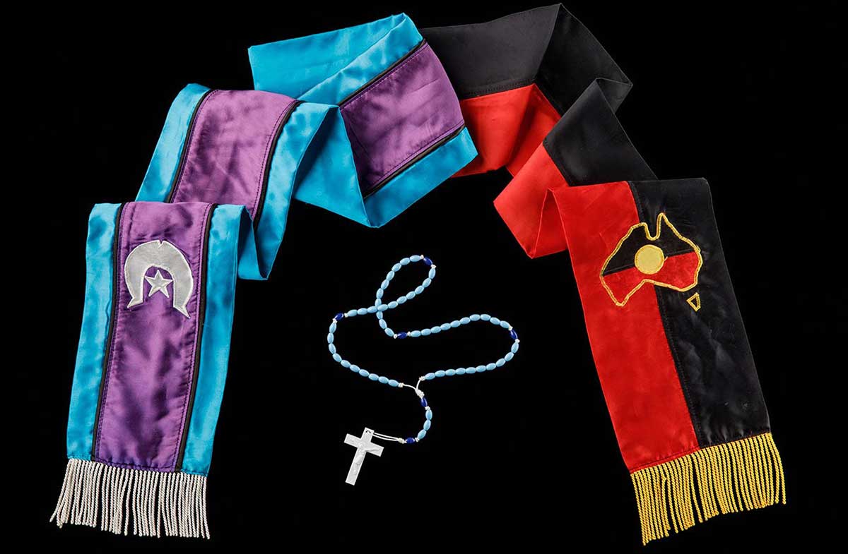 Description A sash that is half red and black and half aqua and purple. The end of the red and black side has yellow fringe along the edge. There is an Aboriginal flag in the shape of Australia abouve the fringe. The purple and aqua side has white fringe with a whiteTorres Straight islander flag. - click to view larger image