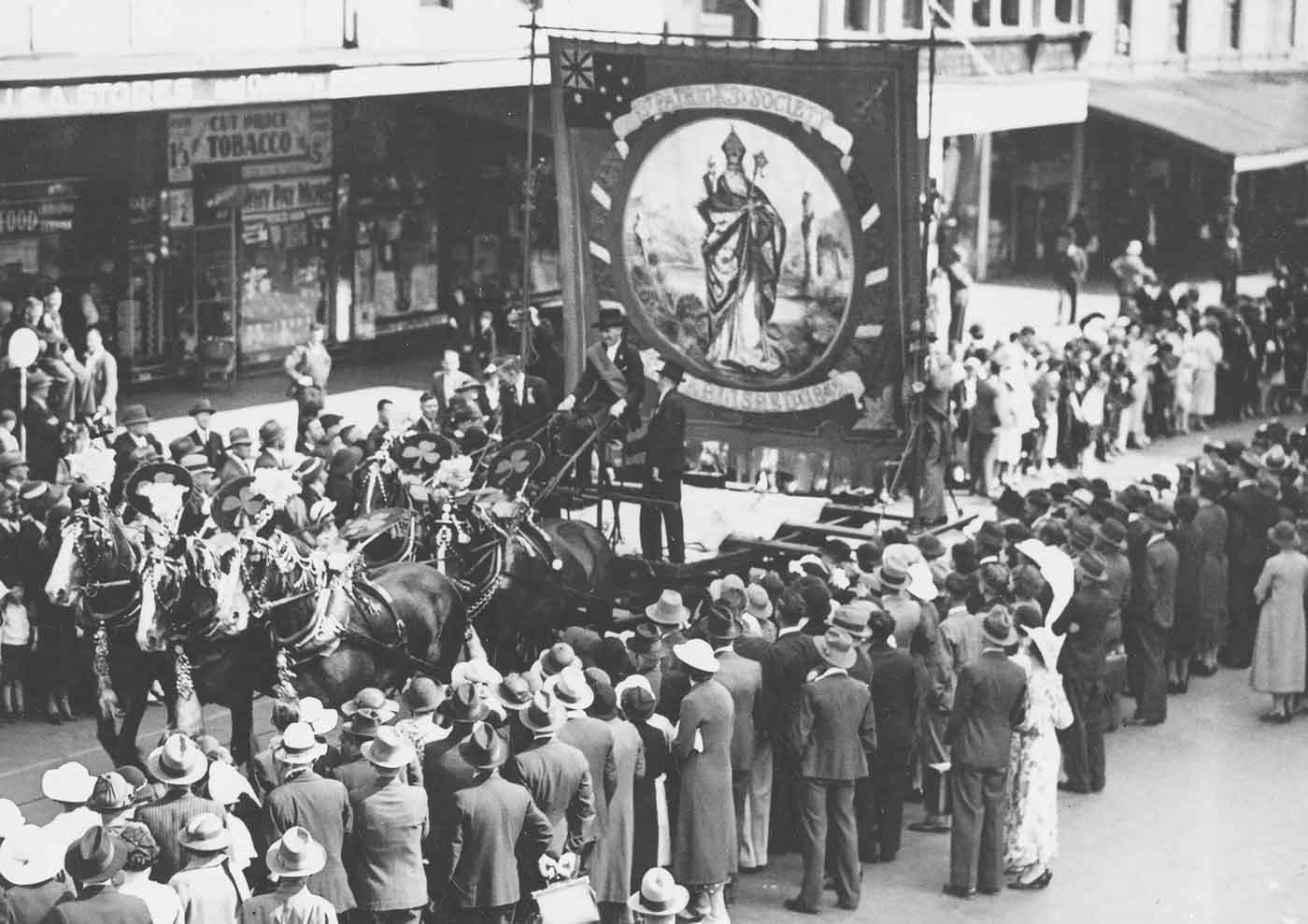 Black and white photo of a procession down a city street featuring a horse drawn float with a large banner, while crowds look on. - click to view larger image