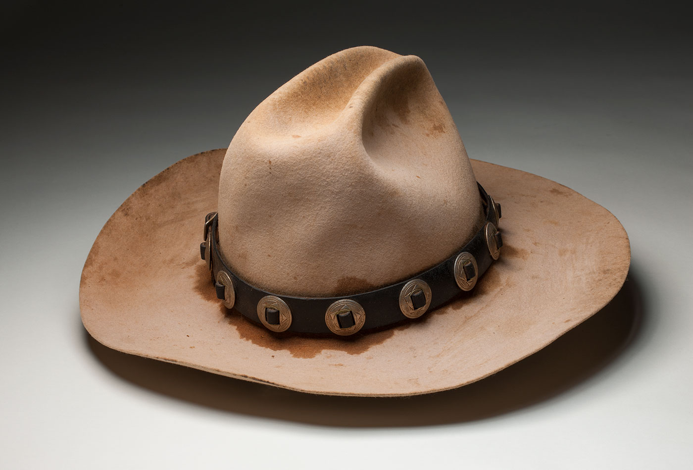 A well-worn light tan coloured hat with a darker leather band, studded with metal.
